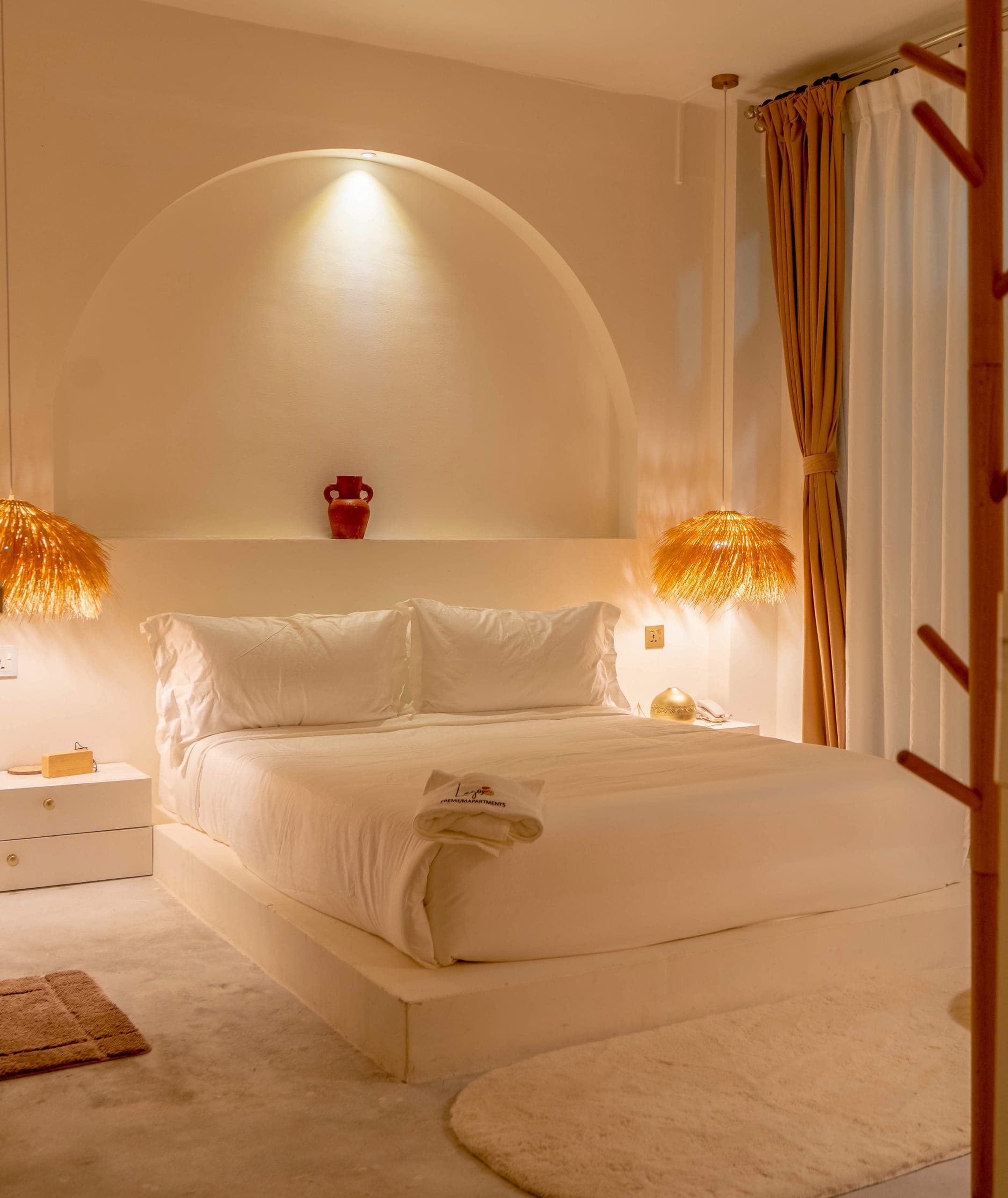 Deluxe Bedroom at Lagom: a white platform bed under a lit plaster arch, straw pendants and linen curtains