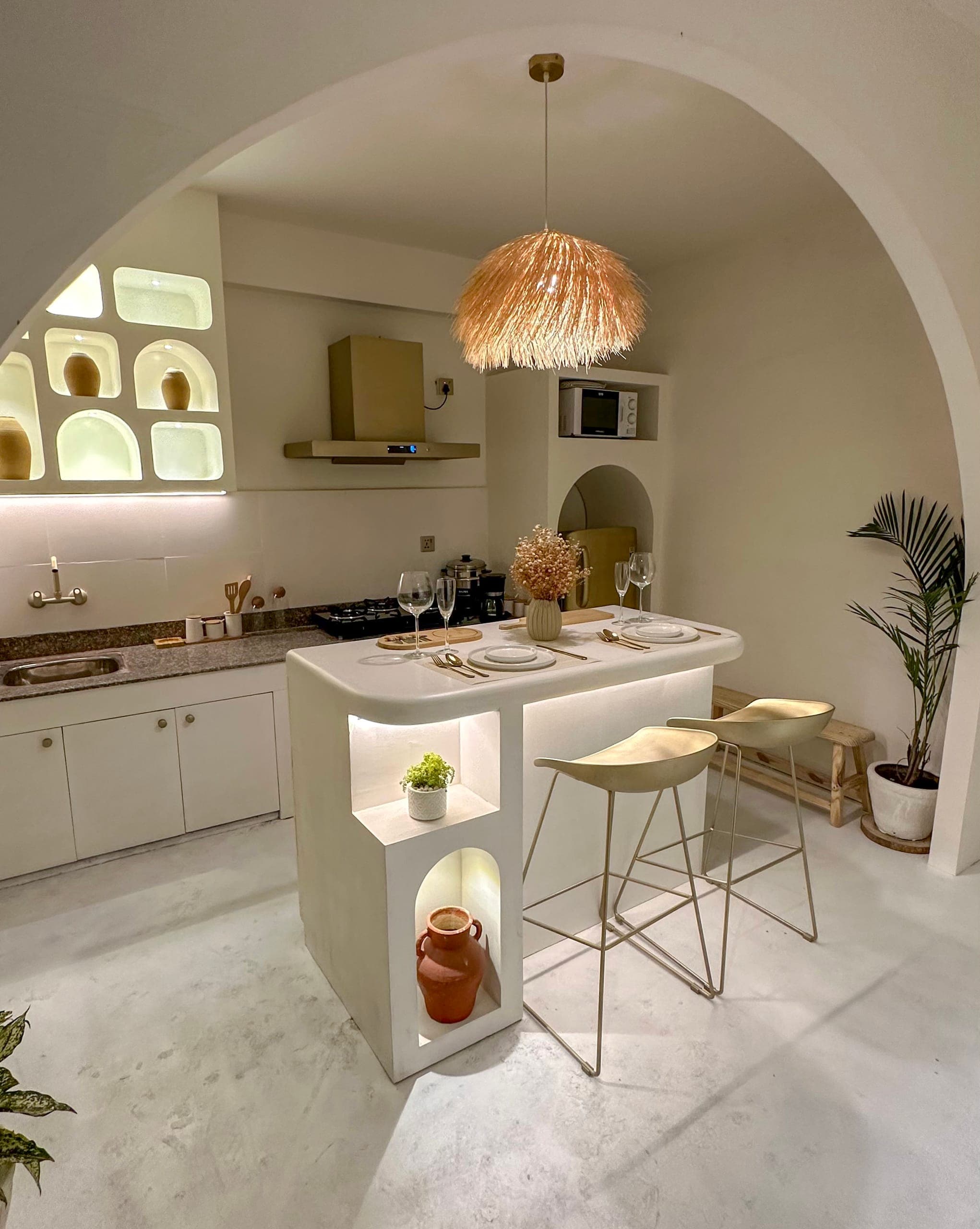 1BK at Lagom seen through a plaster arch: a kitchen island set for two, bar stools, a straw pendant and lit niches