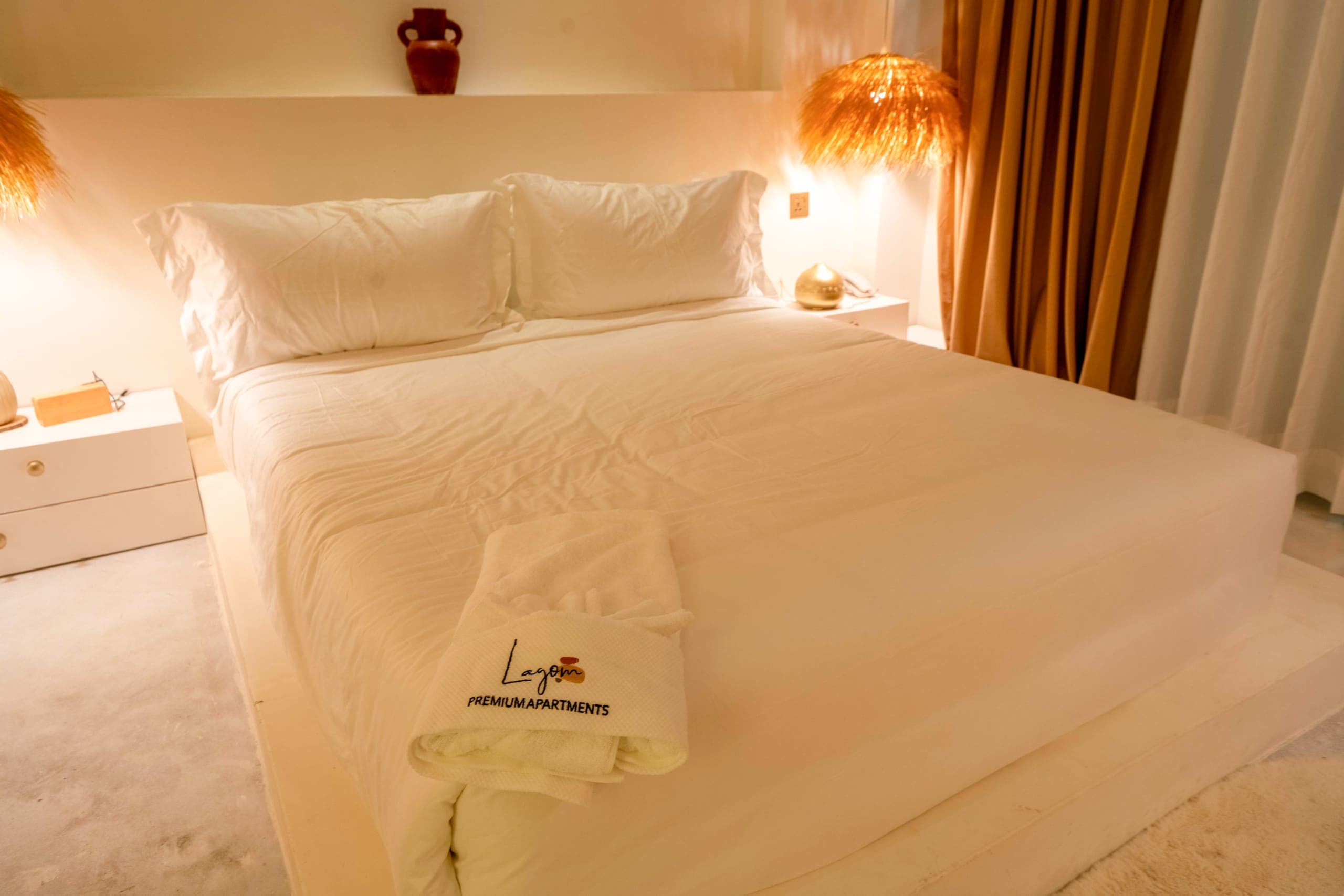 Deluxe bed made up with white linen and a Lagom towel, a straw pendant glowing beside it