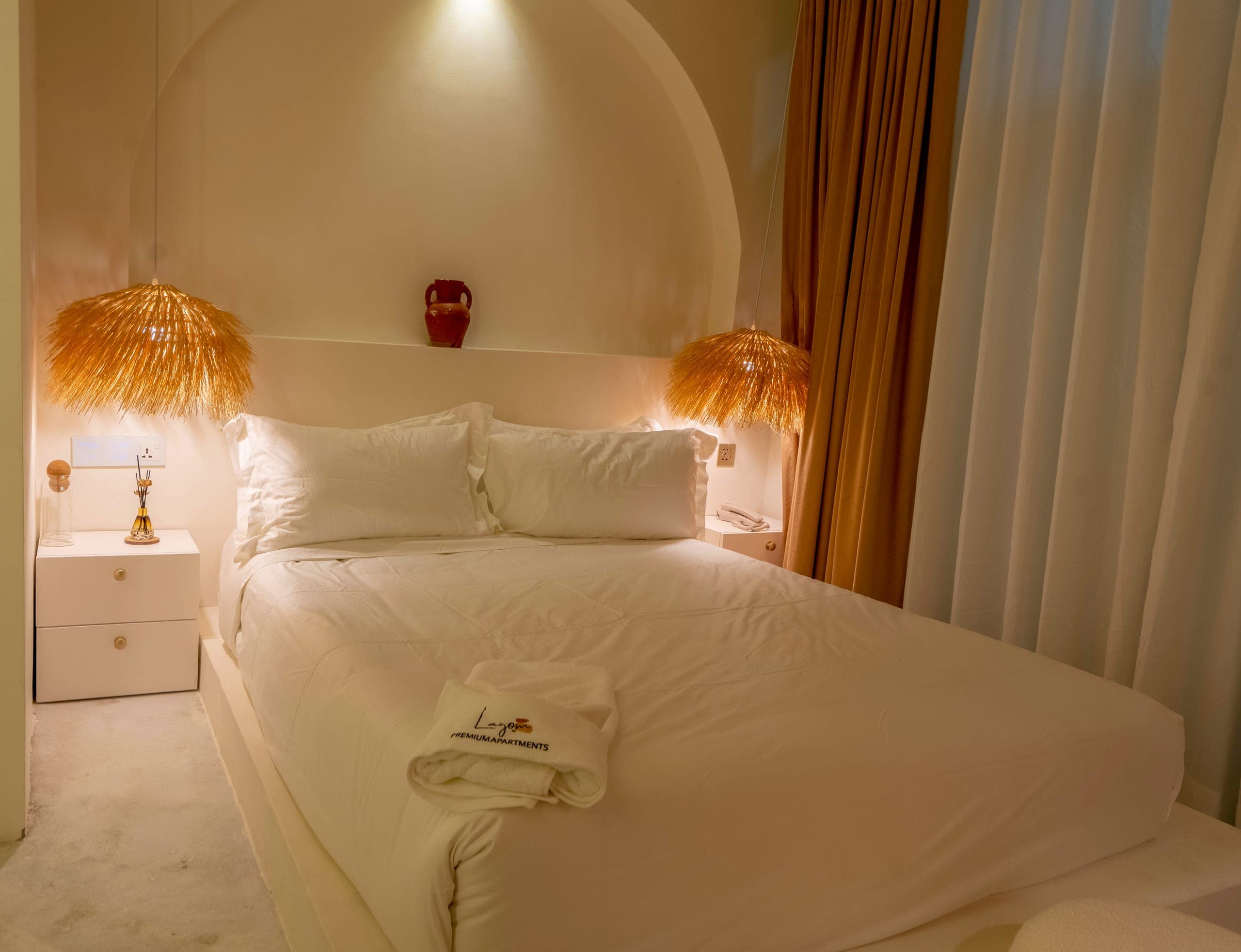 Studio bed under a lit plaster arch with straw pendants, a bedside drawer and linen curtains