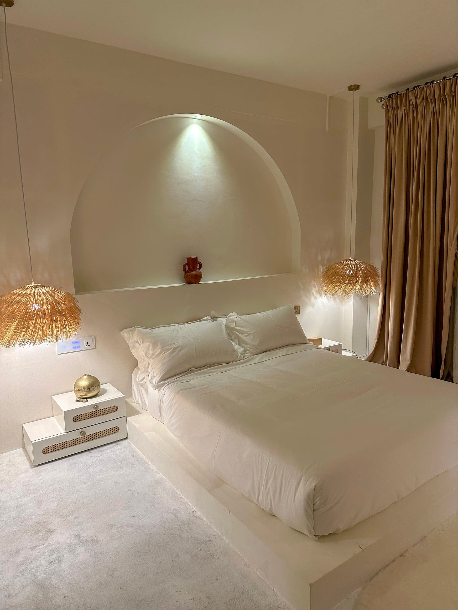 1BK bedroom with a white platform bed under a lit plaster arch, straw pendants and linen curtains