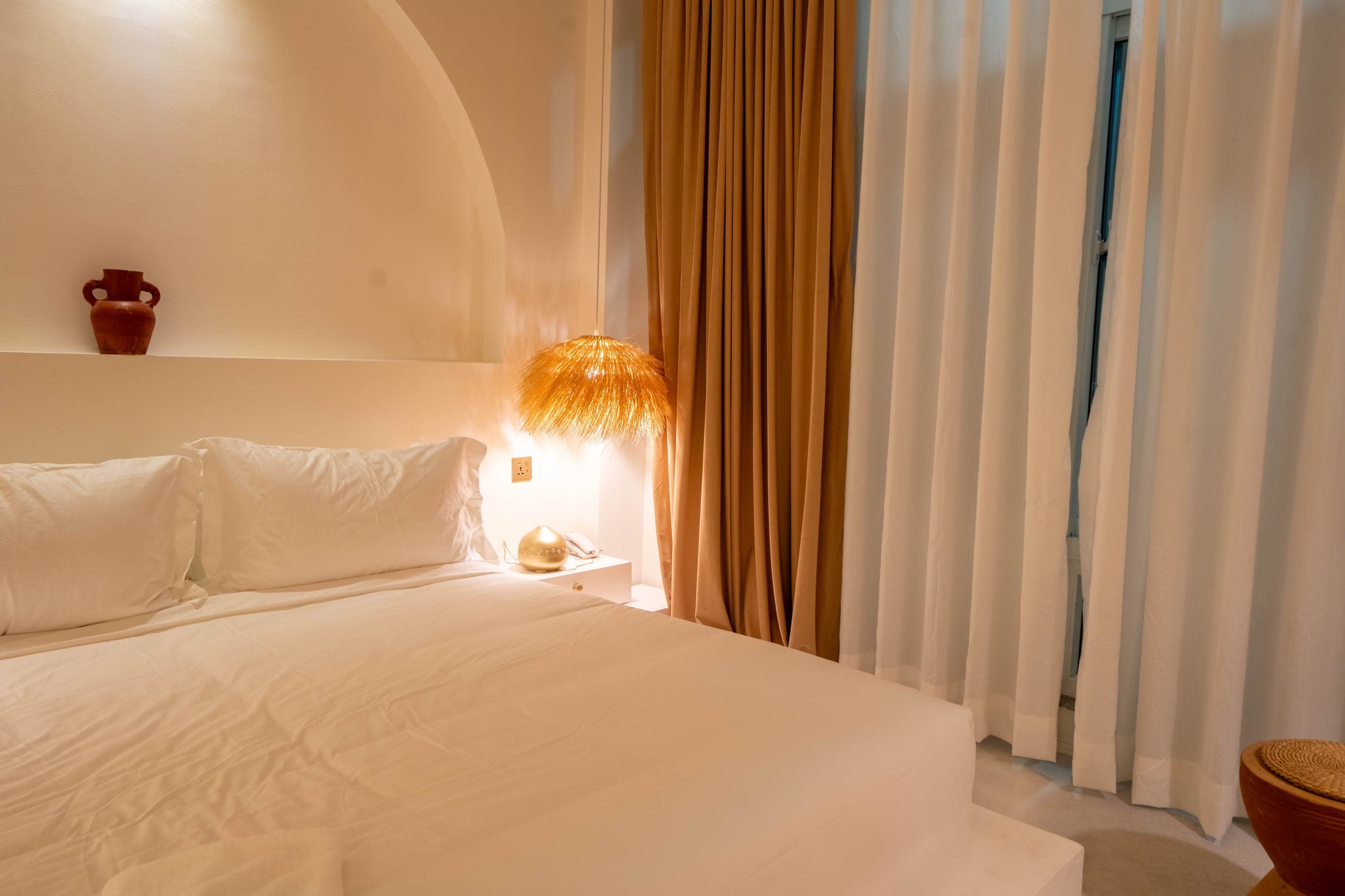 The Deluxe bed beside full-height sheer and linen curtains, a terracotta jug on the arch ledge