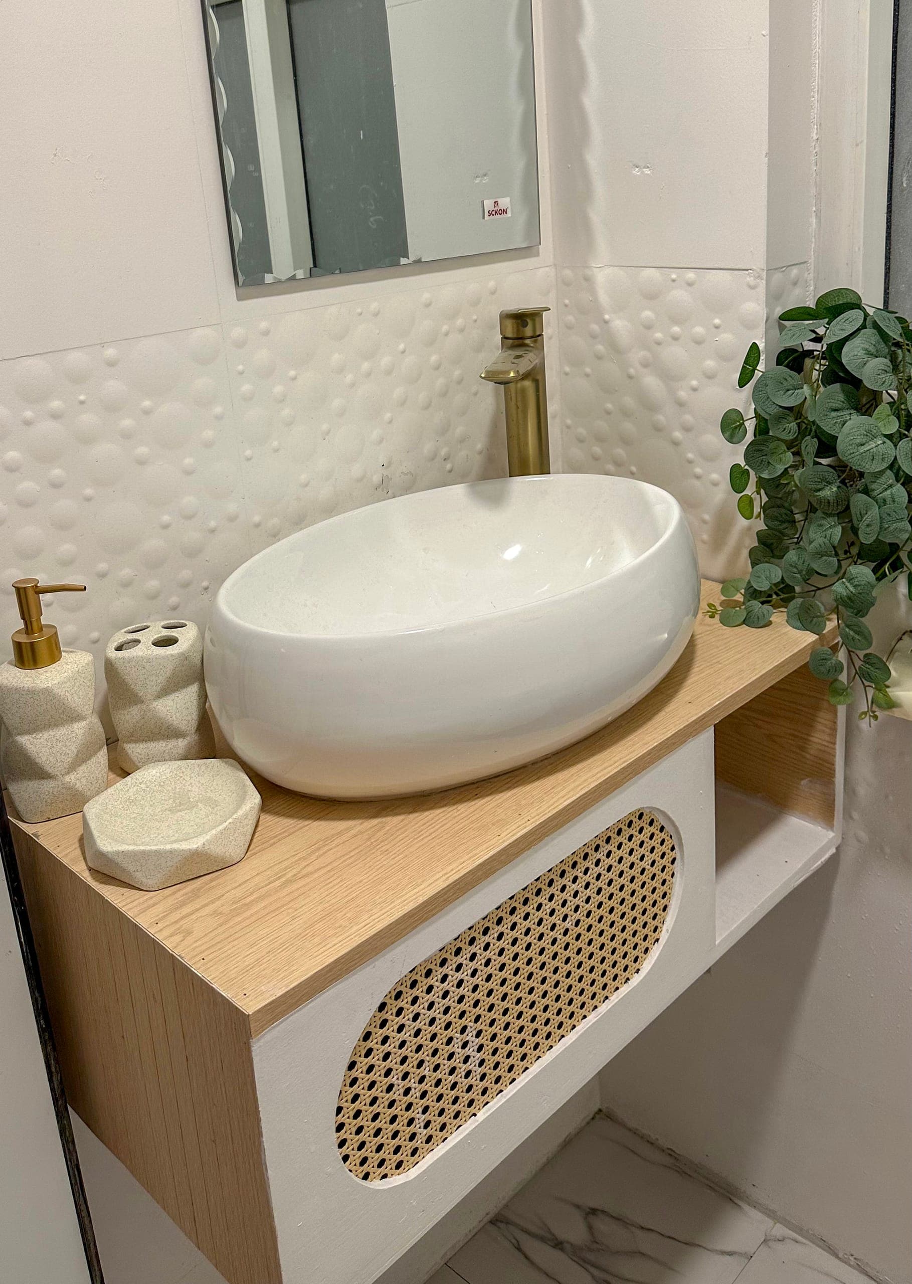 1BK bathroom: an oval vessel basin with a brass tap on a rattan-front vanity and trailing greenery