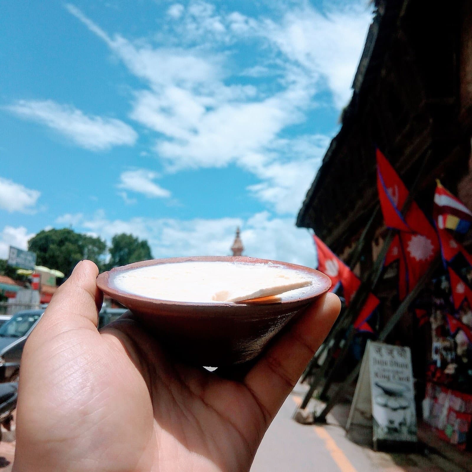 A clay bowl of juju dhau, Bhaktapur's sweet yoghurt, held up against a blue sky and Nepali flags