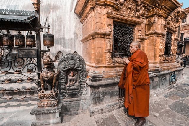 A monk in maroon robes praying at a carved shrine beside a row of prayer wheels