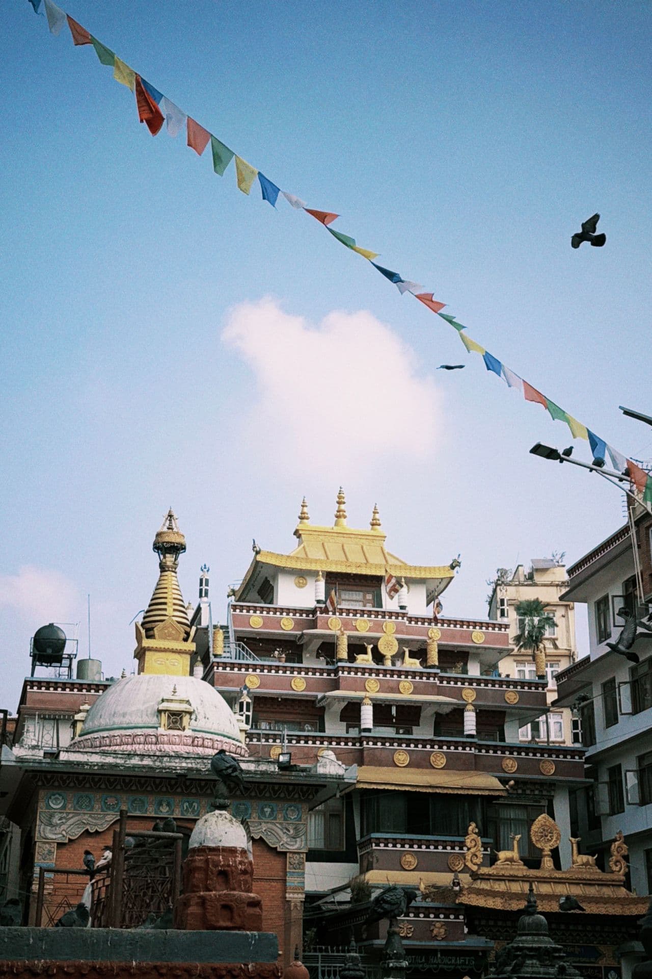 A gilded Tibetan-style monastery rising over a small white stupa, prayer flags strung across a blue sky