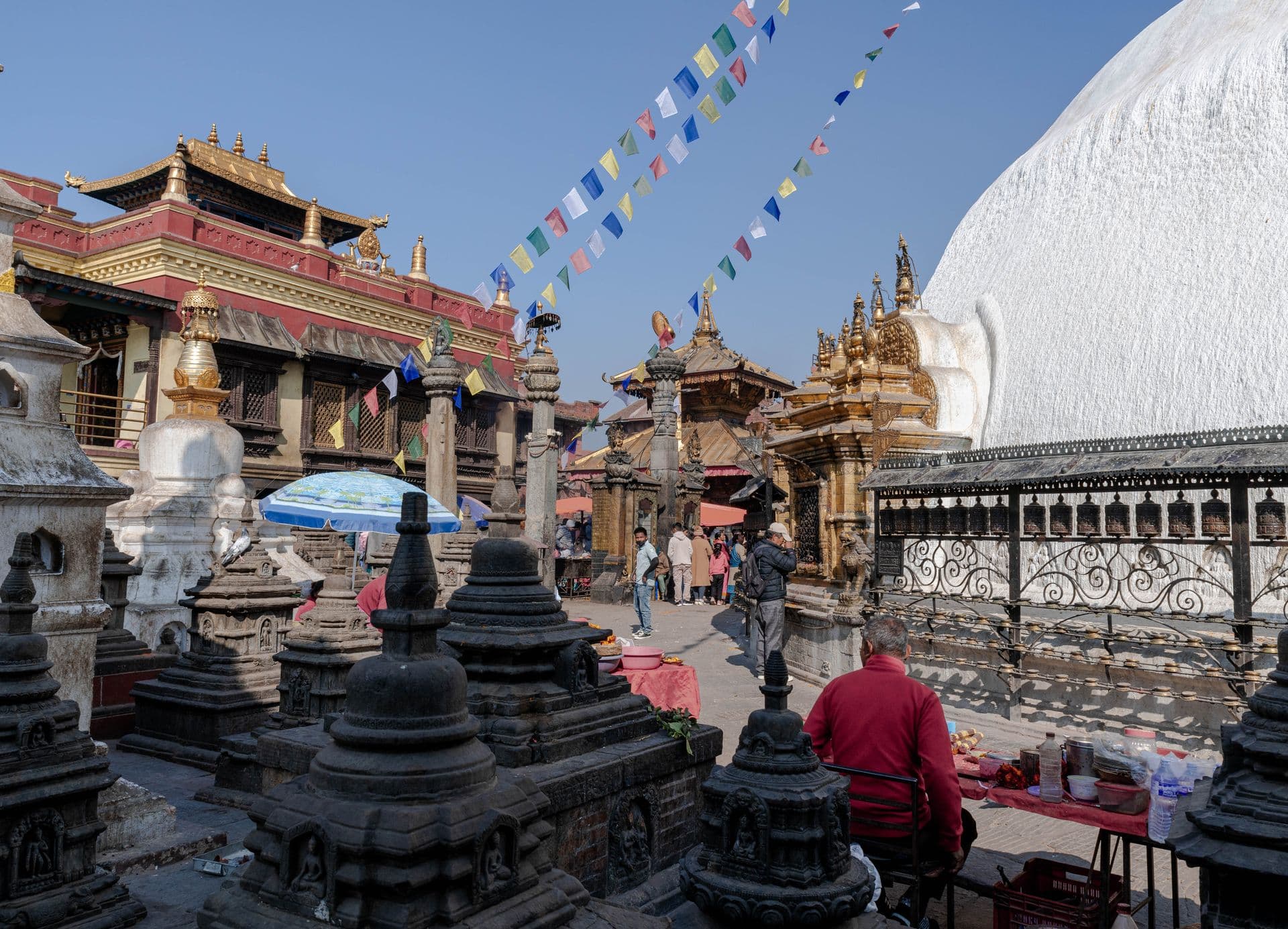 Temple courtyard with black stone shrines, golden pagoda roofs, prayer wheels and a whitewashed stupa dome